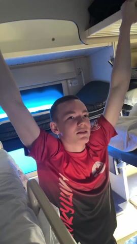 A fair-skinned young man with short brown hair is pulling himself up towards the top bunk of a train bed. He is wearing a red t-shirt with a black fade. The shirt has a circular logo on the chest and an inscription in Cyrillic down the side. The train cabin is small, with two bunk beds and a small window with blue light filtering through. The lower bunk is made with white sheets. The man appears to be struggling slightly as he pulls himself up. There is an additional bunk above the top bunk.