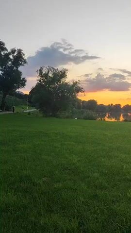 A video shows a sunset over a lake with clouds in the sky. The foreground is a grassy field. There are trees on the far side of the lake. A paved path runs along the lake in the middle ground, and a few people are walking on it. The sun is setting behind trees on the horizon, casting a warm glow on the clouds and water.