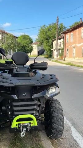A black ATV is parked on the side of a paved road. The ATV is facing forward and is slightly angled towards the left. The front of the ATV is visible, including the handlebars, headlights, and large tires. A bright yellow winch is attached to the front of the ATV. In the background, there is a street with houses on either side. The houses are of various colors and architectural styles. There are trees and power lines visible. The sky is blue with some clouds. The sun appears to be shining, casting shadows. The overall scene appears to be a rural or suburban setting.