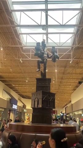 The video shows a large bronze statue in the center of a shopping mall. The statue depicts a man in a dynamic pose, balancing several boxes on his arms and head. He is standing on a tiered base that resembles buildings. The mall has a high, vaulted ceiling with exposed beams and skylights. People are visible in the background, some walking through the mall and others seated on a curved bench in the foreground. The atmosphere appears to be that of a busy, public space.