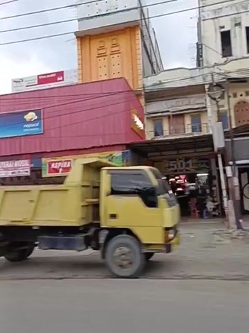The video shows a street scene with several buildings. A yellow dump truck is parked on the side of the road. To the left of the dump truck, a yellow van is also parked. A person is riding a motorcycle past the vehicles. The buildings have various signs, including "VIVA MOBIL" and "ASPIRA". The sky is overcast. The video appears to be shot from a moving vehicle. The scene is likely a commercial area.