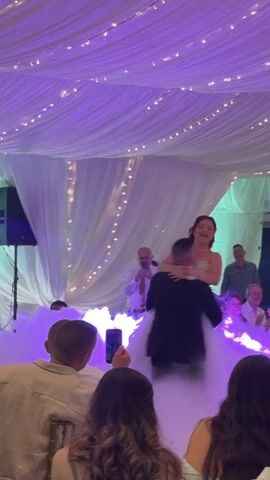A man in a black suit gently lowers a woman in a white wedding gown, who has red hair, onto a smoky, purple-lit dance floor. The man is holding the woman from behind, supporting her waist and back. She is smiling and appears to be laughing. They are surrounded by an audience, some of whom are clapping and smiling. The ceiling is decorated with white draped fabric and string lights. There are also string lights on the white curtains in the background.