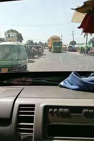 The video, taken from inside the cab of a vehicle, shows a busy street scene in what appears to be a rural or semi-rural area. A large green truck loaded with what looks like hay or sugar cane dominates the view ahead, as it is stationary in the lane. Several people are milling around on the side of the road. There are other vehicles including tuk-tuks and vans. A man in a red shirt is crossing the road in front of the camera vehicle. The dashboard is visible, with a blue knitted item and other objects placed on it. A red cloth ornament hangs from the rearview mirror. The weather appears to be sunny and clear.