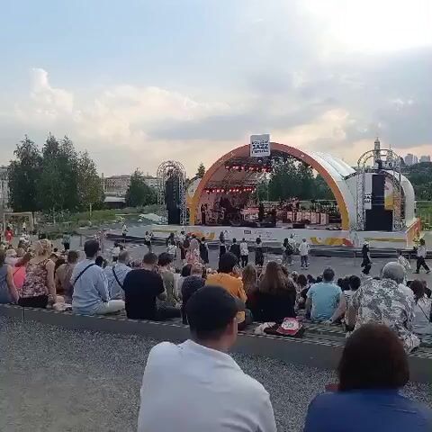 This video shows an outdoor jazz festival with a large audience sitting on benches facing a stage. The stage is covered by a large, arched structure. There are musicians on stage and people are visible in the audience. The sky is cloudy and the lighting suggests it is late afternoon or early evening. The crowd appears to be enjoying the performance.