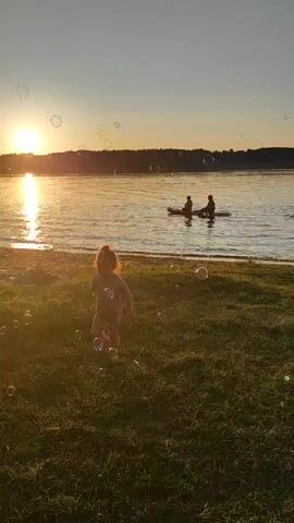 At sunset, a child runs on a grassy shore. Bubbles float in the air and on the grass. In the distance, two people are in a kayak on a lake. More people are in the water near the kayak. The sky is clear and the sun is setting on the left side of the frame, casting a warm glow.