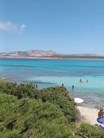 The video shows a tropical beach on a sunny day. The water is clear and turquoise, with some rocky areas visible. Several people are swimming and sunbathing on the beach. There are also some trees and bushes along the shoreline. In the background, there is a body of water with boats and a distant landmass.