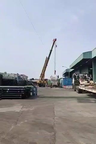 A crane is lifting a large, white, rectangular object. There are several people standing near the object, guiding the lift. The crane is yellow and red, and it is extended high into the air. The scene takes place in an outdoor industrial area, with a paved ground, a building in the background, and stacks of pipes or metal on the left. A truck is parked to the right of the scene.