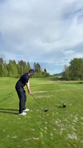 A man wearing a navy blue shirt, dark pants, and a baseball cap is playing golf. He stands on a bright green, grassy golf course under a partly cloudy sky. Behind him, there's a backdrop of green trees, and a glimpse of a distant hill. He swings a golf club, hitting a golf ball that's on a tee. After hitting the ball, he watches its trajectory. A black tee marker sits nearby on the grass.