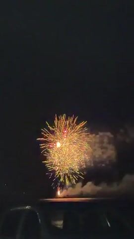 A video shows a firework exploding in the night sky. The firework is yellow and red and creates a burst of light and smoke. The video is taken from a car, with the top of the car visible in the foreground. The sky is dark and clear, with no clouds. The firework is the only source of light in the scene, aside from the faint reflection of the firework on the car's surface.