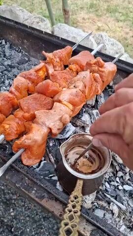 The video shows skewers of marinated raw meat being grilled over hot coals. Next to the grill, a small pot of liquid, likely coffee or a sauce, is being stirred with a spoon. The scene appears to be outdoors, with natural elements such as rocks and greenery visible in the background. A hand is visible reaching into the frame to stir the liquid. The lighting suggests it is daytime.