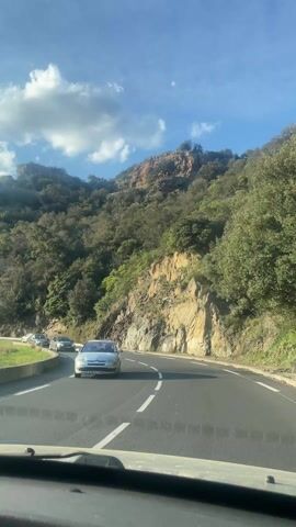 The video shows a drive along a winding road next to a mountain. Cars drive in both directions on the road. The mountain is covered with trees and rocky terrain. The sky is blue with white clouds. The sun is shining through the clouds, creating a bright and sunny atmosphere. The perspective is from inside a car, looking out the front windshield.