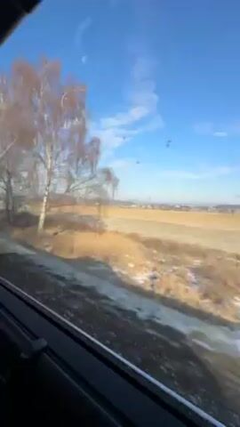 The video shows a landscape view seen from a moving train window. The sky is blue with scattered clouds. Adjacent to the train tracks, there is a field covered with dry vegetation and patches of snow or frost. Bare trees are seen along the edge of the field. The camera moves with the train, causing the scenery to change. A road sign with a red triangle is briefly visible through the train window.