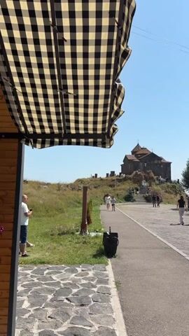 A sunny day unfolds with a checkered canopy in the foreground. A paved path leads towards a hill topped with a stone church. Several people walk along the path and ascend towards the church. Green trees and grass dot the landscape under a clear blue sky. One man stands near the foreground, looking at his phone.