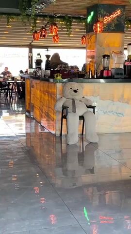 A video shows the interior of a cafe or restaurant with a polished grey tiled floor that reflects overhead lights. In the mid-ground, a large, light-colored teddy bear wearing a dark tie sits on a black chair positioned next to an illuminated bar counter made of translucent, amber-toned stone. Various coffee machines, blenders, and bar equipment are visible on the counter behind the teddy bear. Above the bar, "Espresso" is written in white neon script, alongside green neon accents, and several amber-colored glass globes hang from the ceiling, which is decorated with artificial green vines. To the left, a red Coca-Cola refrigerator is visible. In the background, through large windows, multiple patrons are seated at tables, appearing to dine or socialize. The camera executes a slow, smooth pan to the left, revealing more of the cafe's interior and additional seating areas.