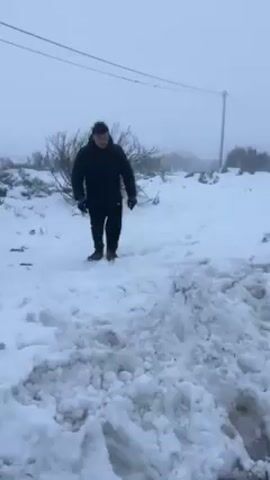 A man in a black jacket, black pants, and brown work boots is running through a snow-covered area, kicking up snow with his right foot. The weather is overcast and foggy.  A utility pole and some bushes are visible in the background.