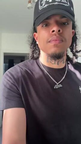 A young man in a black t-shirt, black sweatpants, and a black baseball cap dances in a white kitchen. He has dark skin, long curly hair pulled back, a thin goatee, and several tattoos, including one on his neck. He wears a silver chain with a pendant. The kitchen has white walls, a white refrigerator, and a light fixture with three cylindrical lights. A doorway is visible in the background leading to another room with a window covered by dark curtains. The man adjusts his cap, then begins to dance with a variety of hand and arm movements.