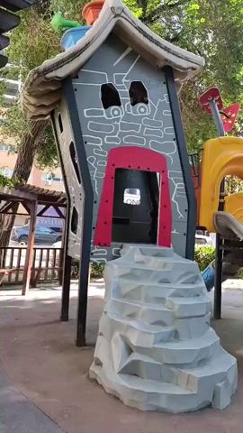 The video shows a playground with a brightly colored play structure. The structure has a gray, cartoonish house with red accents and rock-like steps leading up to a red door. To the right of the house is a yellow slide with a green entrance. The playground is situated outdoors, with trees and buildings visible in the background. The ground is covered in reddish-brown material, likely rubberized playground surfacing. The lighting suggests it is daytime.