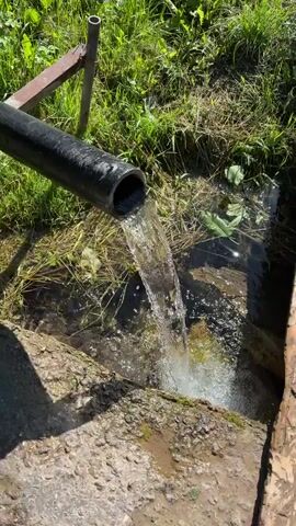 A black pipe is angled downwards, discharging a stream of clear water into a small pool of water. The water splashes as it hits the pool. The area around the pool is overgrown with green grass and dried vegetation. A large piece of wood, possibly a log, is visible in the foreground. A metal support structure is partially visible in the upper left corner of the frame. The scene appears to be outdoors on a sunny day.