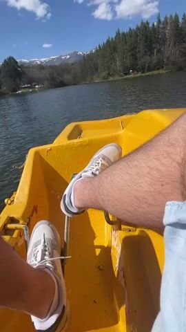 A first-person perspective video shows two people riding a red and yellow paddle boat on a lake. The video starts with a shot of a person's legs pedaling the boat. They are wearing white sneakers and light blue shorts. The camera pans around to show the lake, which is surrounded by trees and mountains. A woman wearing sunglasses, a white hoodie with a blue design, and black leggings is sitting on the boat. She is holding a cell phone. The camera pans around again to show the other person's legs. The sky is blue with some white clouds.