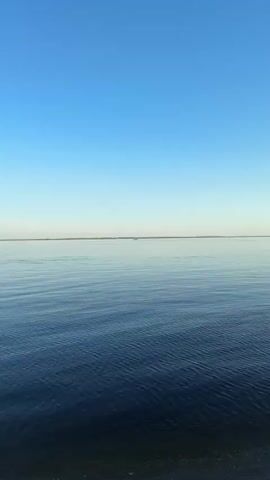 The camera films a wide shot of a calm body of water under a clear blue sky. The water has gentle ripples and reflects the sky's color. In the distance, a thin strip of land is visible on the horizon. The foreground shows a dark shoreline with some visible pebbles or rocks. The lighting is bright and natural. There are no people or objects visible in the water.