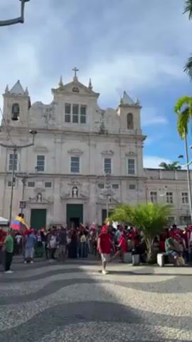 The video shows a group of people gathered in a public square. Many of them are wearing red shirts, and some are holding flags. In the background is a large, ornate church or governmental building. The square has a wave-like pattern on the pavement and is decorated with palm trees. The sky is blue with some clouds. The people appear to be standing around talking, suggesting a gathering or demonstration. The overall atmosphere seems peaceful and potentially political or celebratory.