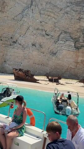 The video shows a view from a boat on a sunny day, with clear turquoise water and a large sandy beach with a shipwreck. Several boats are visible, some carrying passengers. In the foreground, on the boat from which the video is filmed, there are several people. One person is wearing a black hat and a green and black patterned outfit. Another person in the foreground, slightly to the right, is a young boy wearing glasses. Further back on the same boat, another person in a white shirt is visible. In the background, on the beach, is a large, rusted shipwreck. Other smaller boats are near the shipwreck and the beach. The beach is backed by a very large, steep cliff face. The sky is clear and blue.