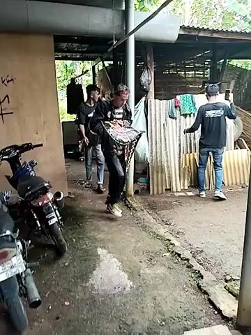 The video shows a group of men in a yard next to a partially open building. Most of the men wear black t-shirts and jeans. One man is wearing black pants and a striped shirt under a jacket. Another man is sitting on the ground, wearing a striped shirt. One man is using his phone to take pictures or video. The setting appears to be a rural or semi-rural area, with trees and vegetation visible in the background. There's also a motorcycle parked nearby. The men appear to be preparing something, possibly related to a cultural event or performance, given the traditional elements in their attire. The ground is a mixture of dirt and gravel.
