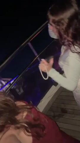 The video shows two women on a balcony at night. The woman in the foreground on the right is wearing a light blue top and is holding a drink. She is pointing to the left. The woman in the foreground on the left is wearing a dark red dress. In the background, a group of people are visible, some of whom are holding drinks. There are lights and what appears to be a DJ booth with flashing lights. The scene suggests a party or club environment. The video is shaky and out of focus in parts.