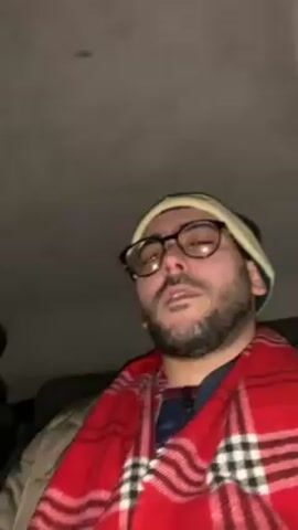 A person with a beard and glasses, wearing a white beanie and a red plaid scarf, is sitting in a vehicle. They are looking upwards and making facial expressions. The interior of the vehicle is dark and the ceiling is visible.