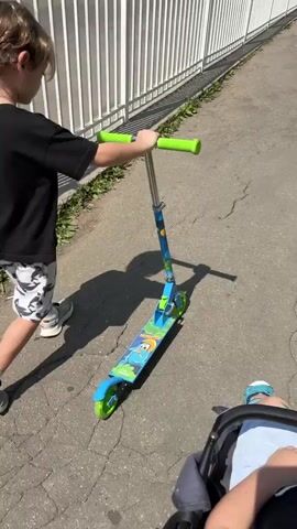 A young boy with brown hair, wearing a black t-shirt and patterned shorts, walks alongside a blue and green scooter with bright green handles. He is holding the scooter by the handlebars. In the foreground, the legs and feet of a baby or toddler wearing blue sandals are visible, sitting in a stroller. The scene takes place outdoors on a paved walkway next to a white metal fence. Sunlight casts shadows on the ground.