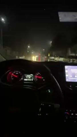 A person is driving a car at night in the rain. The wipers are on. The dashboard is visible with illuminated gauges and a digital display. A smartphone is mounted to the dashboard displaying music information. Streetlights and other vehicles are visible through the windshield.