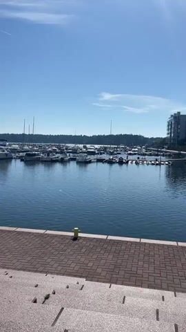 The video shows a marina on a clear, sunny day. Numerous boats of various sizes are docked in the calm, blue water. The marina is lined with a tree-covered shore in the background. A paved walkway runs along the edge of the marina in the foreground. A yellow bollard is visible on the walkway. The scene appears peaceful and idyllic. The sky is a bright, clear blue with a faint contrail from an airplane. The time of day appears to be late morning or early afternoon, judging by the sunlight. The location is Helsinki, Finland.