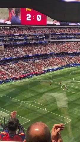 The video shows a wide shot of a soccer stadium packed with spectators. The score displayed on a large screen above the field reads "2:0". There are players on the green soccer field, some in white and some in red uniforms. Spectators are visible in the stands, many wearing red. In the foreground, partially obscuring the view, are the backs of a few spectators' heads, one wearing a red and yellow shirt, and another clapping. A scoreboard overlay indicates a substitution is being made. The overall atmosphere appears to be that of a live sporting event.