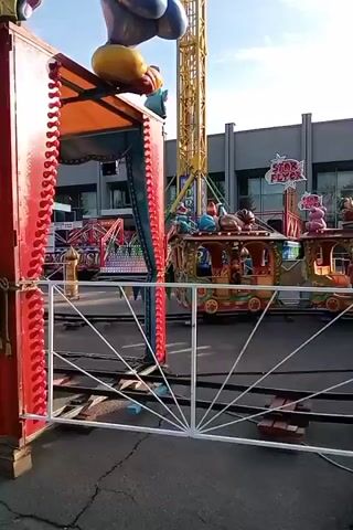 The video shows a children's amusement park ride with a colorful train. A clown character is visible in one of the train cars. In the background, there is a tall tower ride and a building. The sky is blue with some clouds. The ride appears to be stationary at the moment.