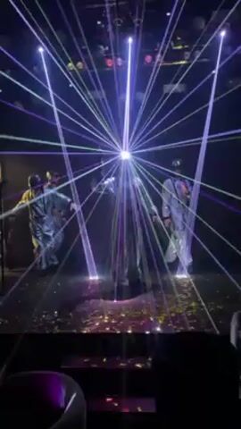 A stage performance featuring four performers in shiny, futuristic costumes takes place amidst a vibrant display of laser lights. The lighting includes a mix of green, purple, and white beams emanating from various points in the ceiling and floor, creating a dynamic and starburst-like effect. The stage floor is littered with confetti, adding to the celebratory atmosphere. The performers are moving, appearing to be dancing or engaging in choreographed movements, illuminated by the swirling light show. Overhead spotlights also contribute to the overall brightness of the scene.