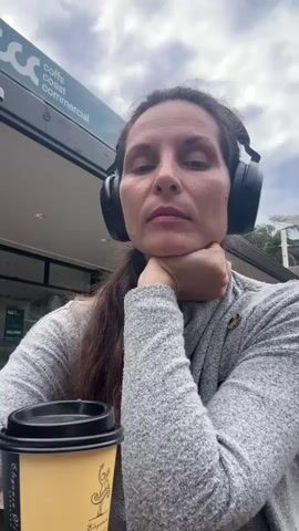 A woman with long, dark hair is sitting outside wearing headphones and a gray sweater. She holds a coffee cup with a tan and yellow design and drinks from it. A sign with white lettering reading "coffs coast commercial" is visible behind her. The sky is cloudy.