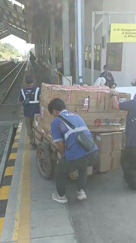 The video shows two men, likely workers, pushing a cart loaded with large cardboard boxes on a train platform. The cart has a wooden frame and two large wheels. The boxes are stacked high, some wrapped in clear plastic. Both men are wearing blue shirts with reflective vests over them. The man on the left is pushing the cart from behind, while the man on the right is pushing from the side. The platform is paved and has yellow and black striped markings along the edge next to the train tracks. There are pillars supporting a roof over the platform. In the background, other people are visible, and a train track stretches into the distance. The scene appears to be a transportation hub, likely a train station, where cargo is being moved.