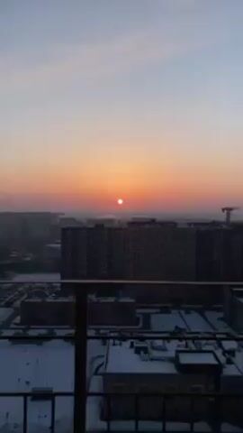 The video shows a winter sunrise over a cityscape. The sun appears as a bright, round orb in the sky, set against a gradient of orange and pink hues. Below the horizon line are several tall, modern apartment buildings with dark exteriors, viewed from an elevated perspective, possibly a balcony. Snow covers the rooftops of lower buildings and parking lots, and there is a visible layer of haze or fog. A railing is partially visible in the foreground. The video is taken from a stationary position. A construction crane is seen on the horizon.