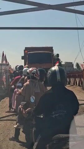 A group of motorcyclists are in traffic. A large orange dump truck is in front of them. The motorcyclists are waiting in line. The sky is clear and blue. The scene is outdoors. There are some flags on the left side of the image. There are some buildings in the background on the right side of the image.