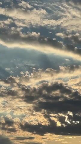 This is a vertical, close-up video of a dramatic sky filled with clouds. The time of day appears to be either sunrise or sunset, given the warm, golden light illuminating the edges of the clouds and casting long, visible sunbeams through gaps. The clouds themselves are varied, with some appearing light and wispy, catching the sunlight, while others are darker and more substantial, creating a strong contrast and sense of depth. The overall mood is serene and awe-inspiring, capturing the beauty of natural light phenomena. There are no people, objects, or specific location cues visible, other than what can be inferred from the sky itself. No text is present in the video. The scene is consistent with a sky view, likely from an open area in Pojarevo, Bulgaria.