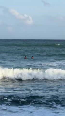 Two people are in the ocean, bobbing their heads above the water. They appear to be surfers, and they are spaced apart from each other. The water is choppy, and there are waves breaking in the foreground. The sky is overcast with some clouds visible. The horizon is a thin line separating the sea and sky.