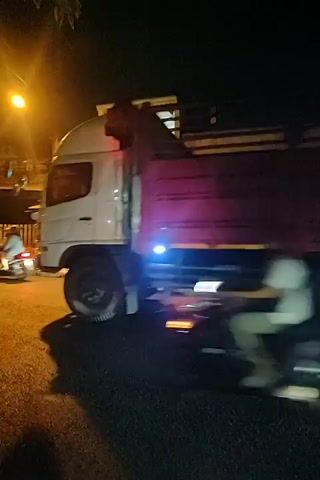 A short video clip shows a night scene on a road. A large truck is moving from left to right across the frame. The truck has a red cargo container and multiple wheels. The truck is illuminated by streetlights and its own lights, including blue lights on the side and red taillights. A person on a motorcycle is also moving from left to right in the foreground, blurring due to motion. The motorcycle has a red taillight. Trees and a building are visible in the background. The lighting is dim, with bright streetlights creating lens flares.
