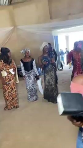A group of four women are dancing in a room, likely at a celebration. Each is wearing a unique, colorful patterned dress. The woman in the middle is holding a white plastic bag. They are all smiling and appear to be enjoying themselves. In the background, more people are visible, and some decorations are present. The camera is held by a person who is partially visible in the foreground. The lighting is bright, and the atmosphere appears festive.