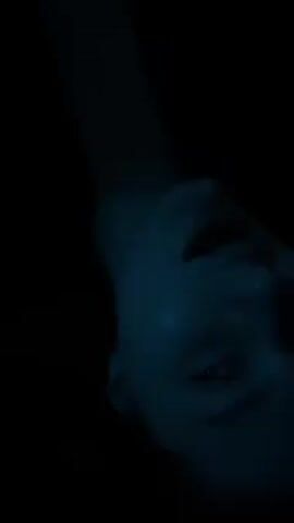 The video shows a close-up shot of a person’s face, viewed from below. The person is lying down, and the lighting is dark with a bluish tint. The face is fairly expressionless, and the eyes are partially open. The background is entirely black.