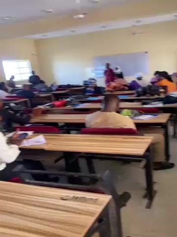 The video shows a classroom environment, likely in a school or university setting. Several students are present, seated at individual desks arranged in rows. Most students are engaged in activities like writing or reading, presumably related to a class. There's a projector screen at the front of the room, suggesting a lecture or presentation might be in progress. The room is well-lit with large windows. The color scheme is neutral, with cream-colored walls. An air conditioner is visible near the ceiling. The floor is tiled. As the camera moves, more students are revealed, some standing or moving around the room.