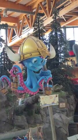 The video shows a large, blue, cartoonish octopus character wearing a golden Viking helmet with horns. The octopus has pink tentacles, each holding a small wooden bucket. Water is pouring out of these buckets, creating a cascading effect. The octopus is positioned above a rocky landscape that appears to be part of an indoor water park or amusement area. Tall, artificial evergreen trees are visible behind the octopus, and the ceiling features exposed wooden beams and industrial lighting. In the background, there are hints of other attractions, including a tall, dark object that resembles a cactus or a stylized hat. The overall environment suggests a playful, themed attraction.
