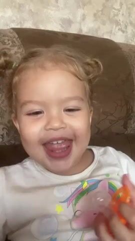 A young child with blonde hair in two buns is smiling widely while holding an orange object up to her mouth. The child is wearing a white t-shirt. The background appears to be a patterned couch.