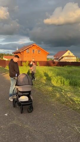 A low-angle outdoor shot shows two men and a toddler next to a baby stroller on a narrow road with grass on the right. The man closest to the camera is pushing the gray baby stroller with a black frame. The second man holds the toddler dressed in a leather jacket and a tan hat. The men and the stroller face the camera as they walk down the road. Behind them is a tan wooden house with a brown fence, another house, and a cloudy, dramatic sky in the background.