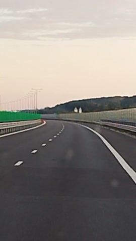 This media file captures a perspective from a vehicle driving on a highway in Merișani, Romania. The scene depicts an empty asphalt road with white dashed lane markers and a solid white line on the right edge. On the left side of the road, a vibrant green sound barrier runs parallel to the highway, with a series of tall lamp posts visible behind it. To the right, a similar, but less prominent, fence-like barrier is in place. Beyond the barriers, a dark, tree-covered hillside forms the horizon. The sky above is a soft, pale orange and pink, with scattered clouds, suggesting either dawn or dusk. Two small, white structures, possibly houses or buildings, are barely visible on the hillside. The overall impression is one of solitude and smooth travel, with no other vehicles or people immediately apparent. The lighting and muted colors indicate a peaceful, perhaps early morning or late evening, atmosphere. There is no visible text in the media file.