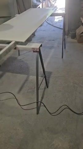 A person is spray painting a long, flat white object that is resting on sawhorses. The object appears to be a door or a large panel. The spray painting is being done in a workshop or garage environment, with concrete floors and some clutter in the background. The person is wearing dark pants and a dark shirt. A black cord is visible on the floor, connected to the spray painting equipment. The lighting suggests it is daytime, with sunlight coming in from an opening in the background.